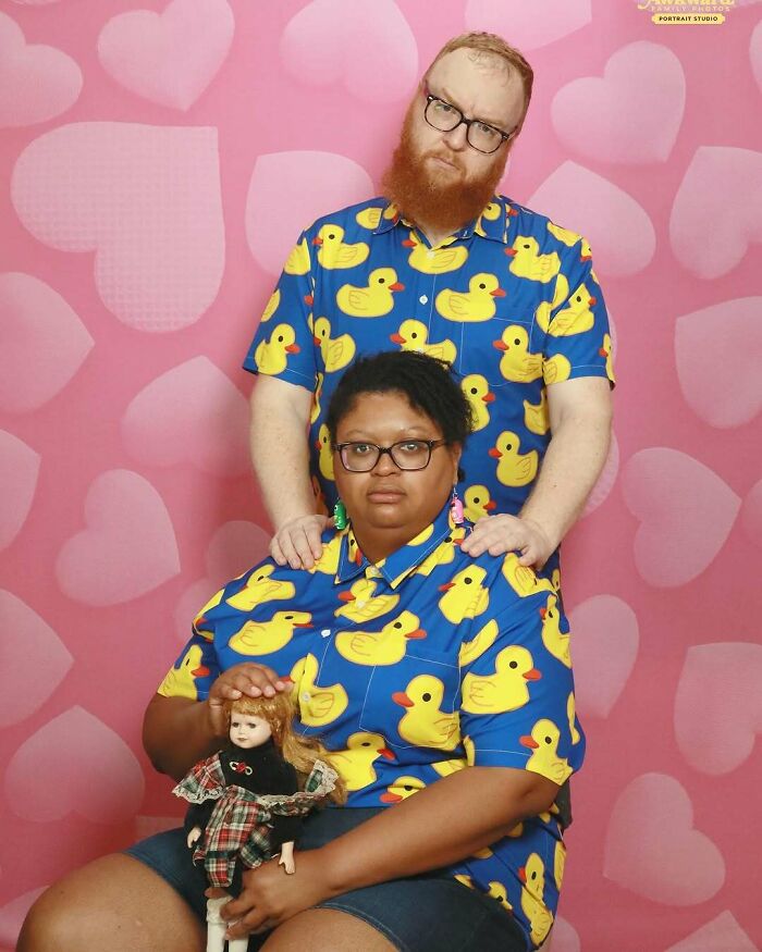 Couple wearing matching duck print shirts posing awkwardly in a studio with a pink heart background and holding a doll.