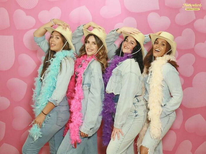 Four women in denim outfits and colorful feather boas posing awkwardly in a studio with a pink heart background, awkward studio pics.