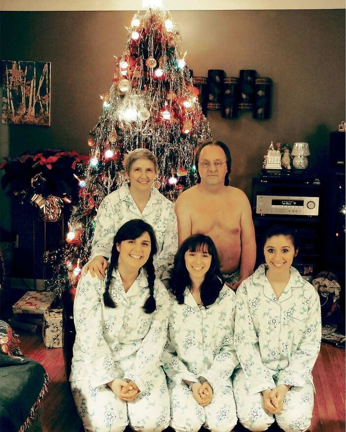 Family in matching pajamas posing awkwardly in front of a decorated Christmas tree, a shirtless man standing behind them.