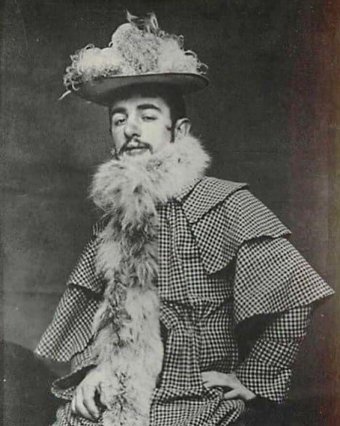 Victorian era man posing in an elaborate checked outfit with a feathered hat and a fluffy scarf in a weird vintage photo.