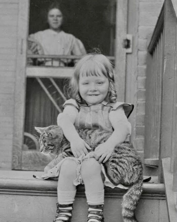 Victorian era photo of a smiling child sitting on steps holding a large tabby cat with a woman in the background.