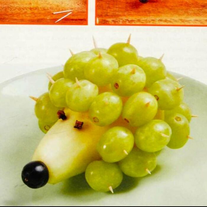 Creative vintage recipe art showing a hedgehog made with pear, grapes, and cloves on a white plate.