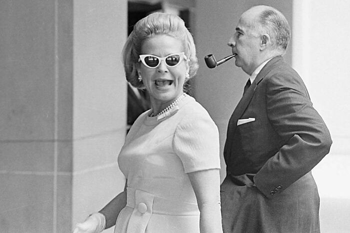 Woman in vintage dress and cat-eye sunglasses smiling, with man smoking pipe, representing overlooked women in history.