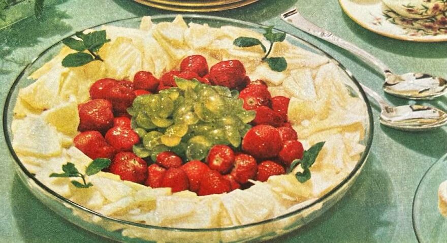 Vintage recipes dessert bowl with strawberries, green grapes, and pineapple slices on a green tablecloth with vintage dishes.