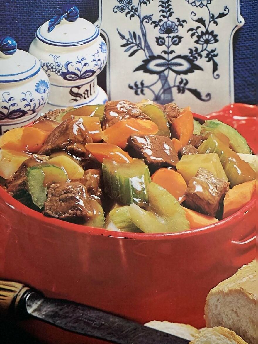 Vintage recipes stew with beef, carrots, and celery served in a rustic red pot alongside salt shakers and bread slices.