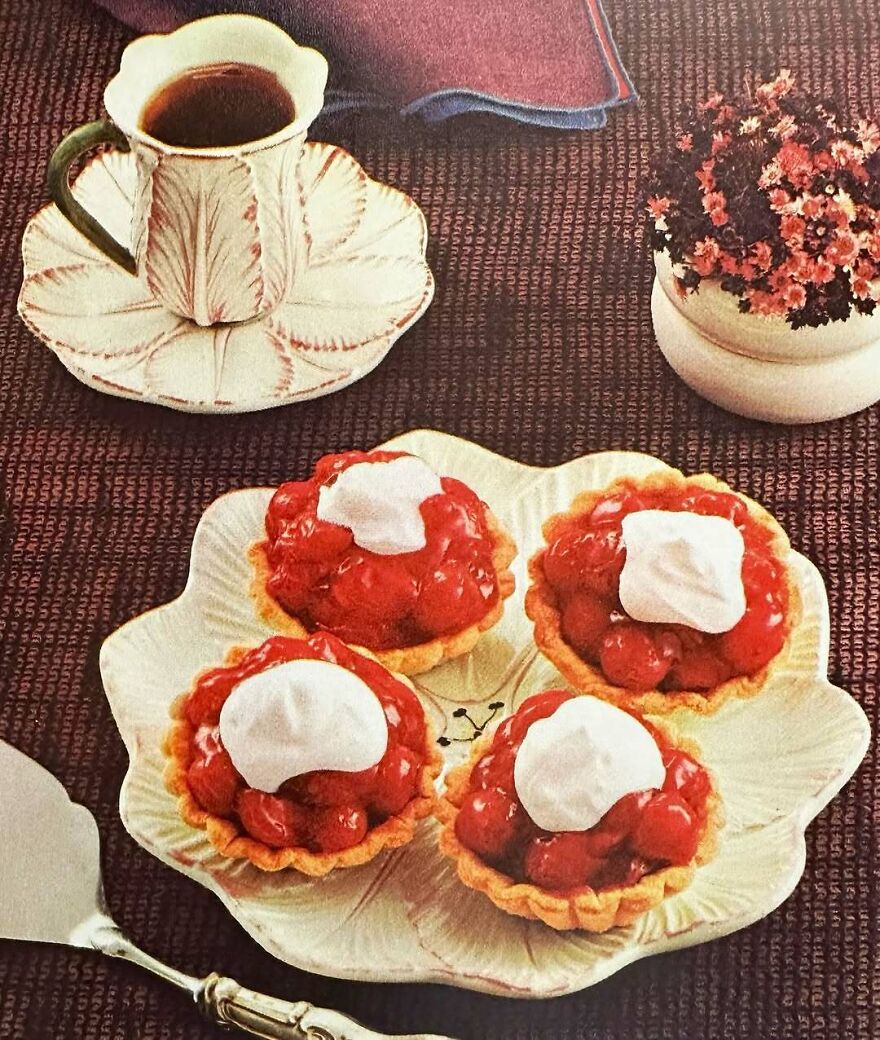 Vintage cherry tarts with whipped cream on a leaf-shaped plate beside a floral cup of coffee and small flowerpot.