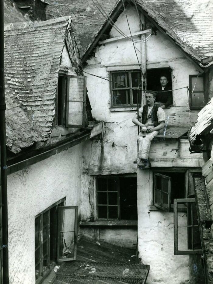 Black and white Victorian era photo showing two people at the windows of an old, worn building with open shutters.