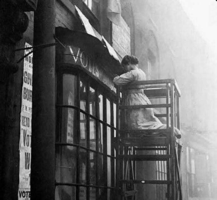 Woman on a scaffold painting a "Votes for Women" sign in a historic black and white photo representing overlooked women in history.