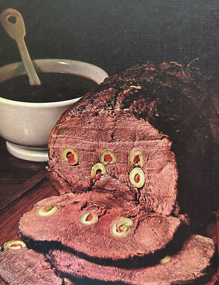 Sliced vintage meatloaf with olives served on a wooden board with a bowl of dark sauce, showing bizarre vintage recipes.