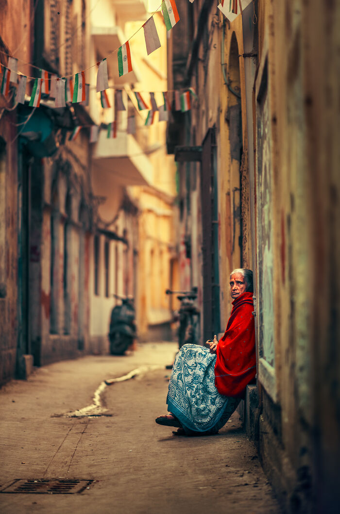 Elderly woman sitting in narrow alley wrapped in red shawl, captured in street photography around the world.