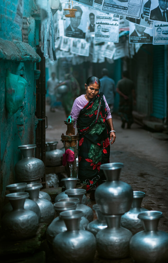 Woman in traditional clothing filling water pots from a street water pump, captured in vibrant street photography shot.