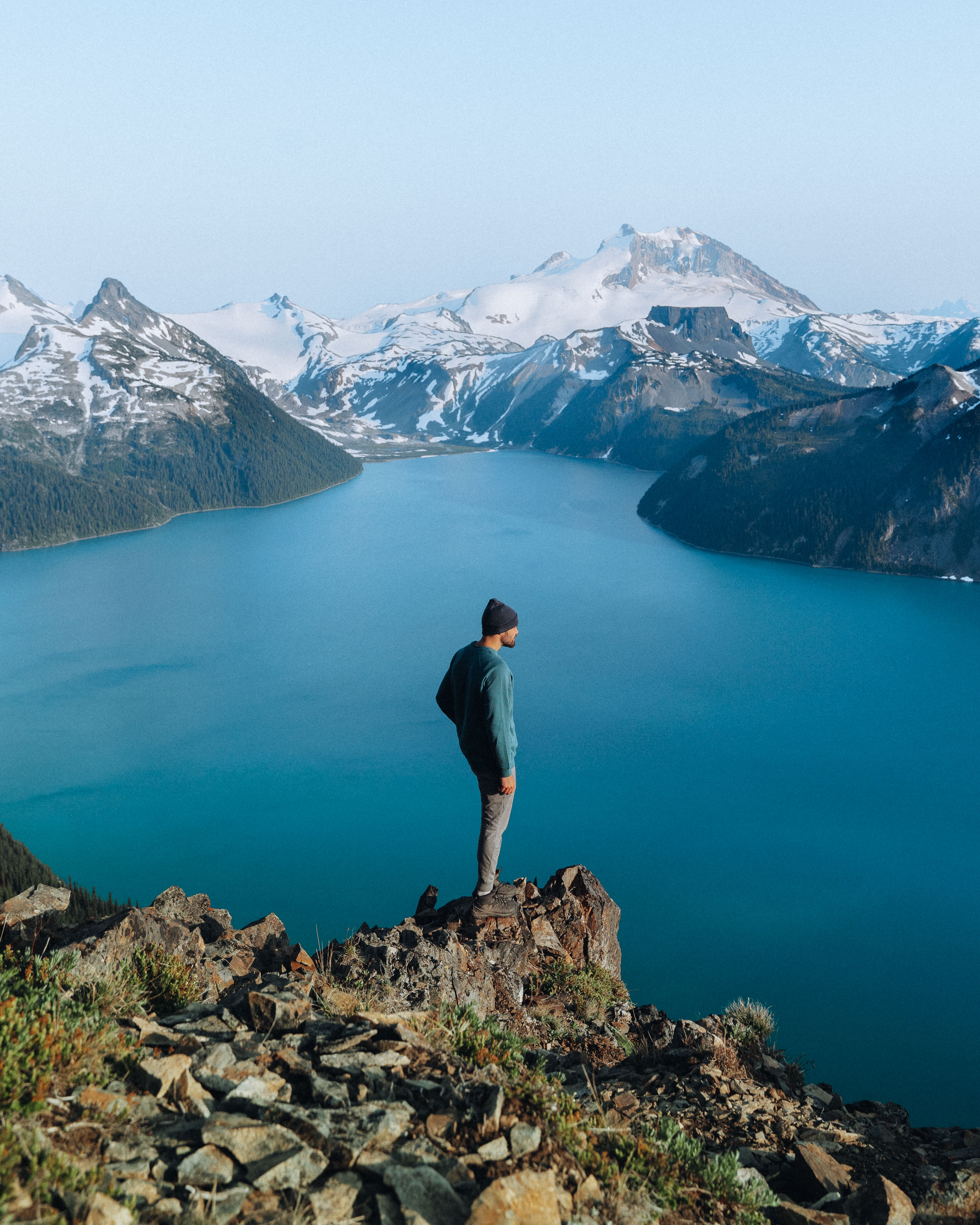 Man standing on rocky cliff overlooking a vast lake and snow-covered mountains, reflecting on purpose and journey.