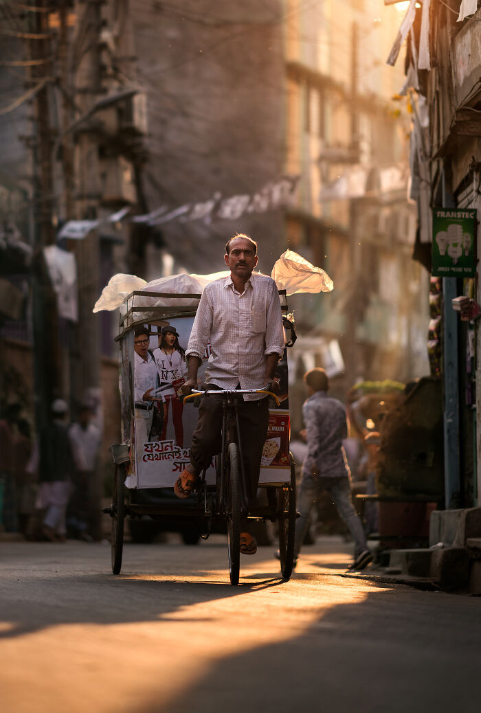 Man riding a bicycle rickshaw in a sunlit narrow street, captured in vibrant street photography around the world.