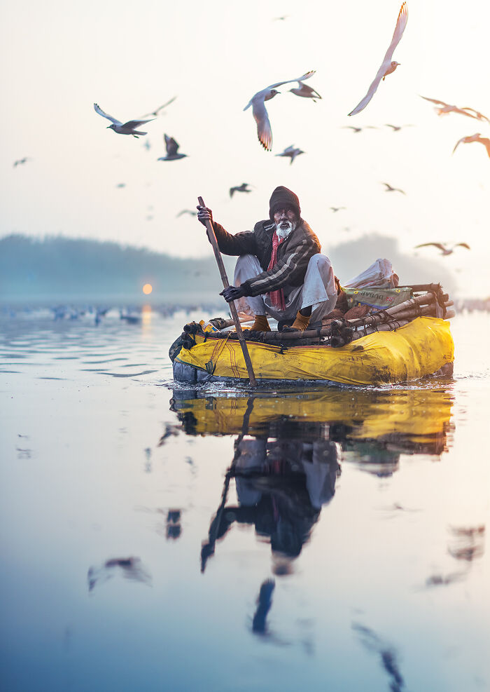 Elderly man paddling a small boat on calm water with seagulls flying overhead in a street photography shot.