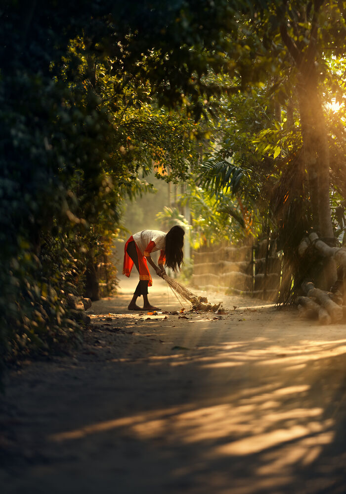 Woman sweeping leaves on a sunlit village path surrounded by lush trees, captured in street photography around the world.