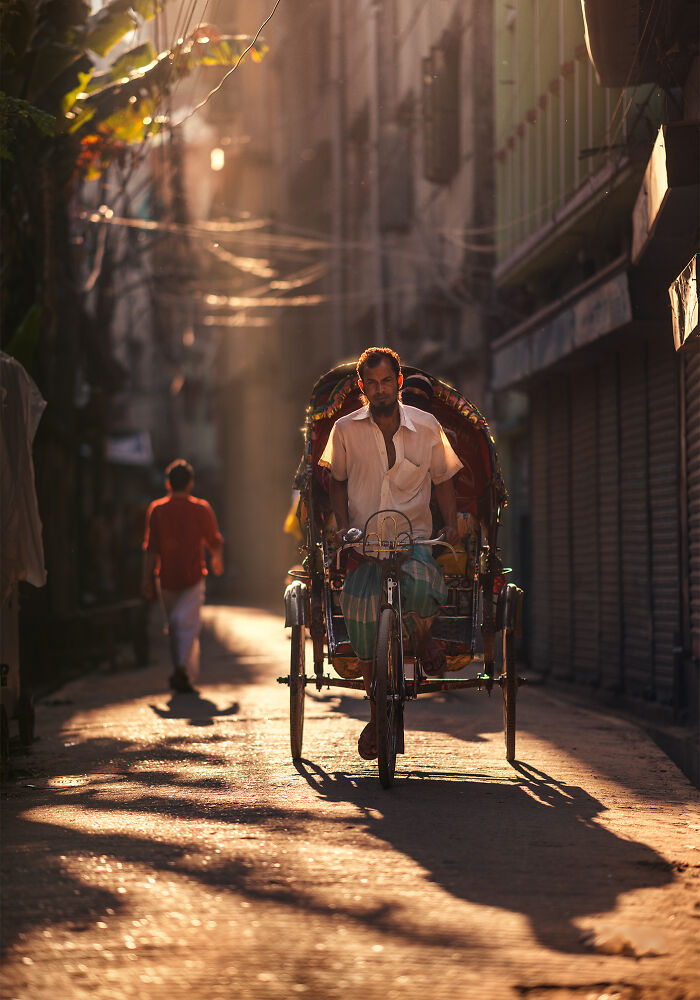 Man riding a cycle rickshaw on a sunlit street with another pedestrian walking in the background in street photography.