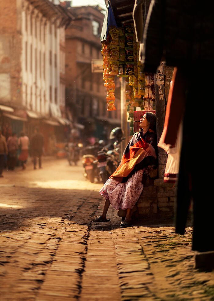 Woman wrapped in a blanket sitting on a street edge in warm sunlight, captured in street photography around the world.