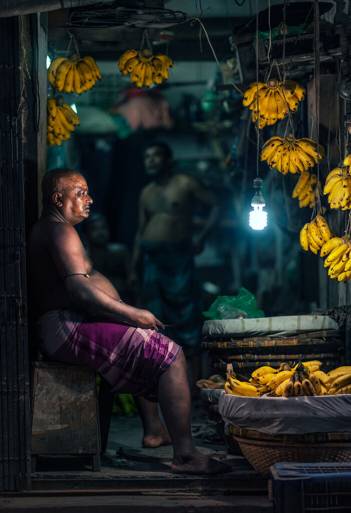 Street photography of a vendor sitting in a dimly lit market surrounded by hanging bunches of bananas.