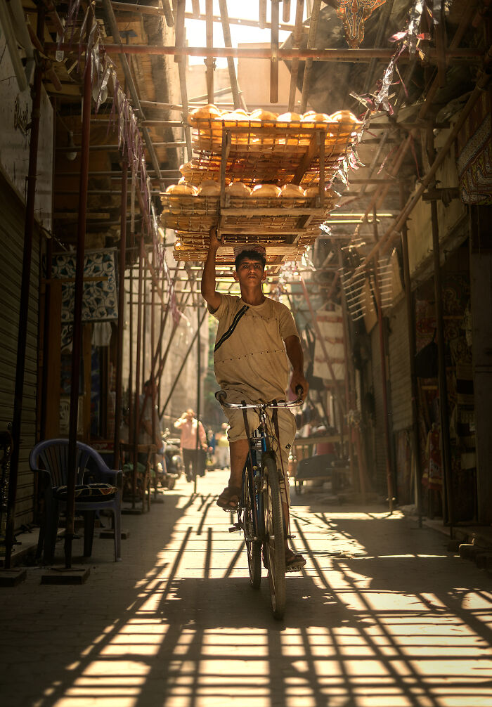 Street photography capturing a young man balancing bread trays on his head while riding a bicycle in a sunlit alley.
