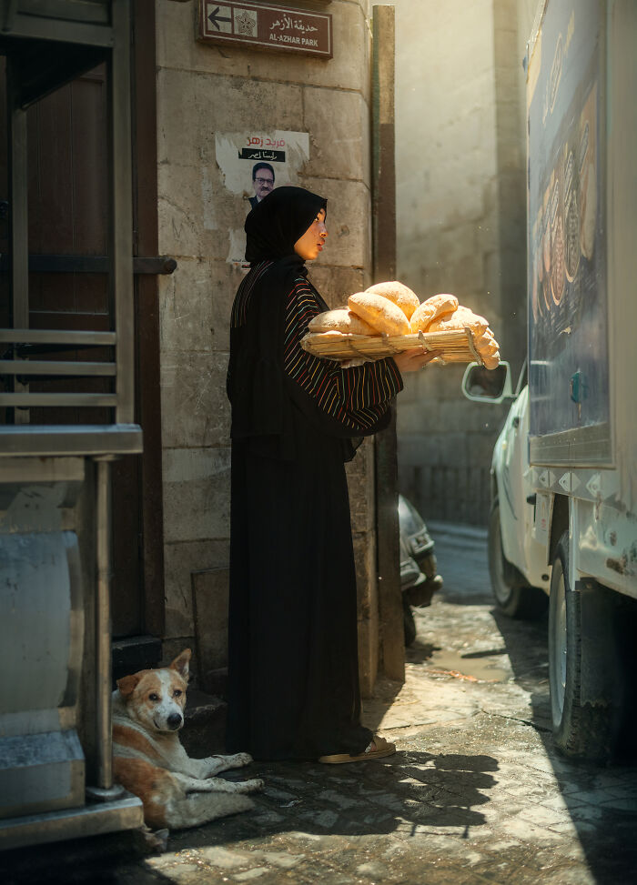 Woman in traditional attire holding bread on a street with a dog nearby, a candid street photography moment.