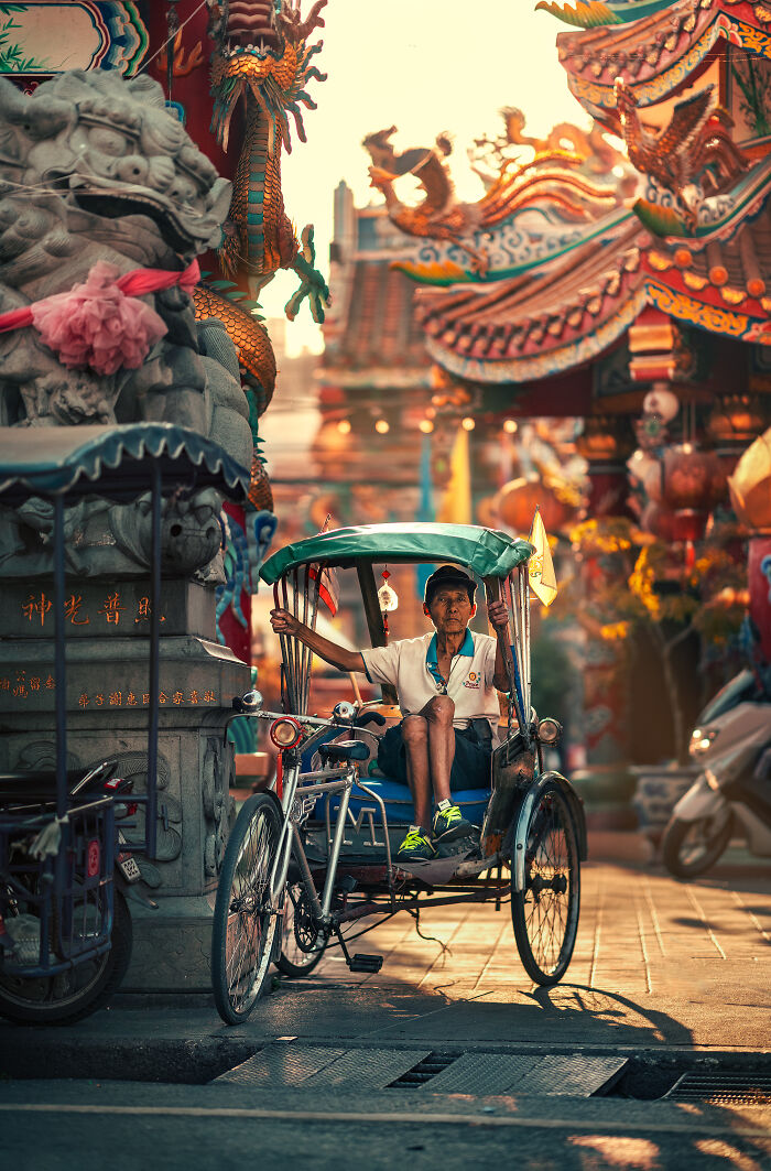 Street photography of a man sitting on a rickshaw near ornate temple carvings at sunset in an Asian city.
