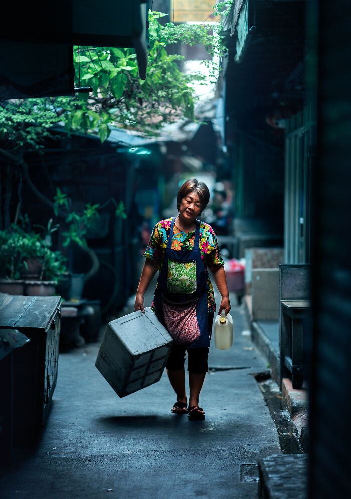 Woman carrying a box and bottle walking through a narrow alley, captured in street photography around the world.