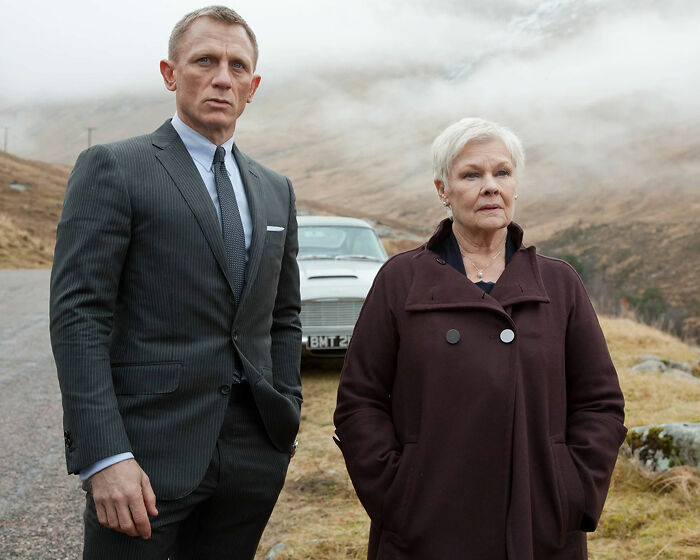 Dame Judi Dench and Daniel Craig standing outdoors near a car, highlighting Judi Dench's health confession about recognition issues.