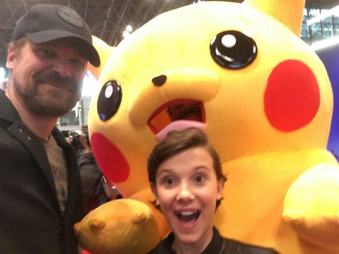 Millie Bobby Brown and David Harbour posing happily with a large Pikachu mascot at a crowded event. Millie Bobby Brown and David Harbour posing happily with a large Pikachu mascot at a crowded event.