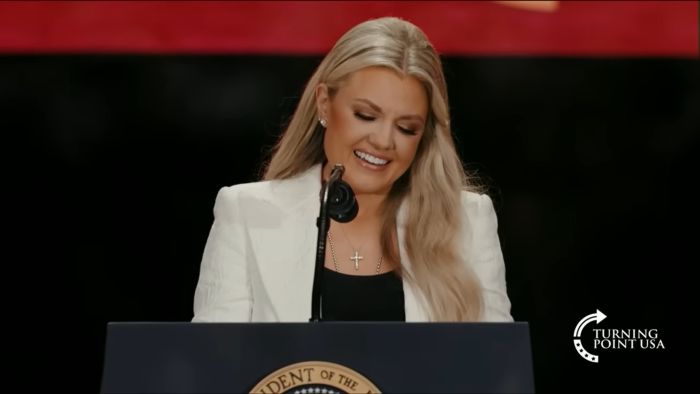 Erika Kirk speaking at a podium wearing a white blazer, involved in country singer touchy-feely hug controversy.