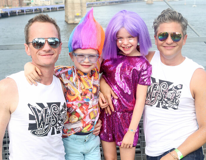 Neil Patrick Harris' daughter Harper in a vibrant purple outfit and wig, smiling with family by a waterfront.