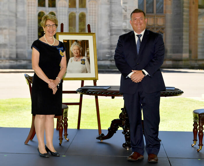 Two formally dressed individuals standing beside a table with a framed portrait outdoors, related to politician resigns news.