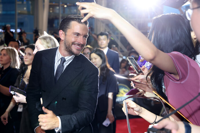 Jonathan Bailey smiling in a black suit interacting with fans at a crowded event under bright lights.