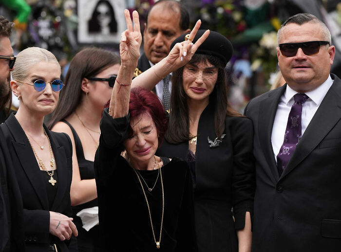Sharon Osbourne emotional and teary at event, raising hand, surrounded by people in black attire and sunglasses.