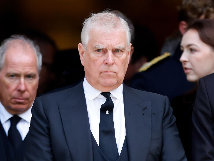 Prince Andrew in a black suit and tie appearing somber, with focus on female touch influencing Andrew&rsquo;s title removal.