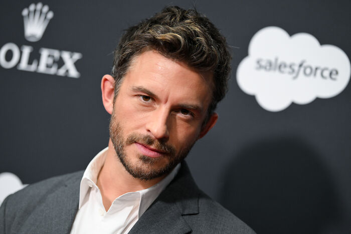 Jonathan Bailey posing at an event, wearing a grey suit jacket, with logos of Rolex and Salesforce in the background.