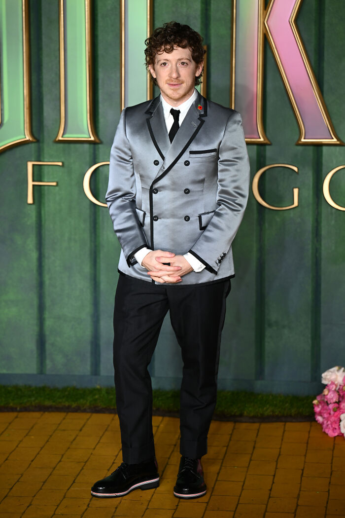 Man in shiny gray double-breasted blazer and black trousers posing at Wicked For Good London premiere red carpet event.