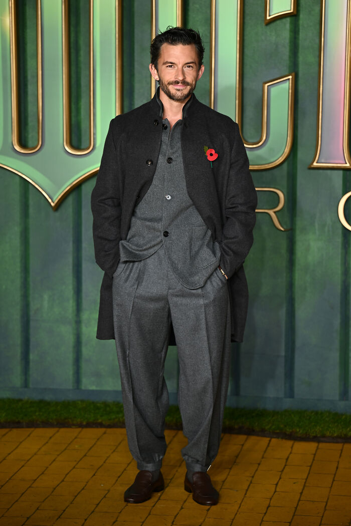 Man wearing a dark coat and gray suit posing on the red carpet at the Wicked For Good London premiere star outfits event.