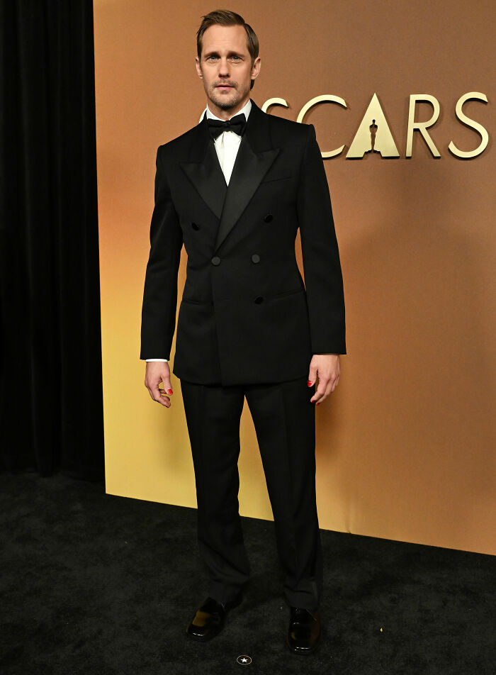 Alexander Skarsg&aring;rd at Oscars event, wearing a classic black tuxedo with a unique red nail detail on one hand.