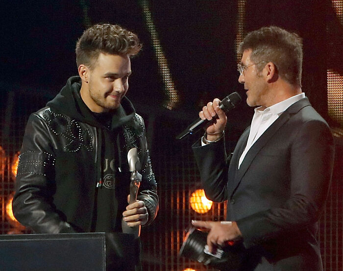 Simon Cowell speaking into a microphone while Liam Payne listens during a public event on stage.