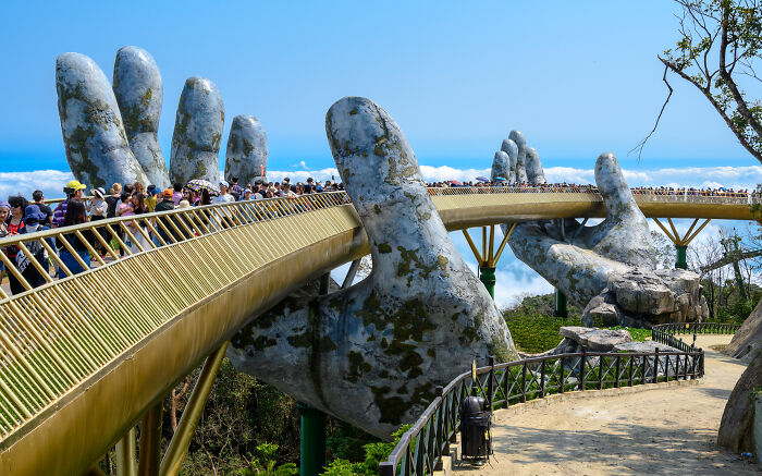 Bridge supported by giant stone hands with tourists walking across, showcasing spectacular sculptures travel destination.