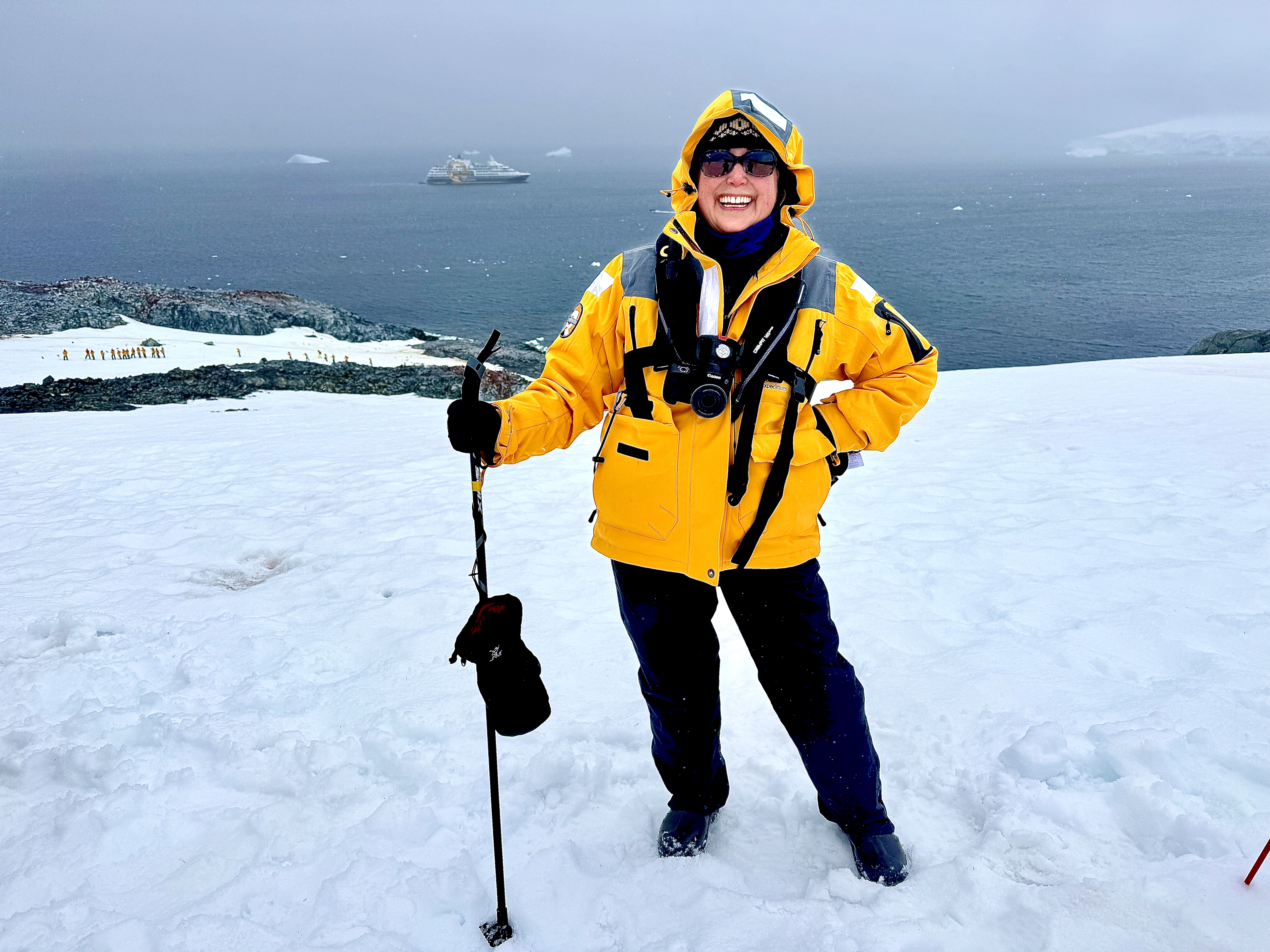 Smiling woman in yellow jacket and sunglasses standing on snowy Antarctica landscape with ocean and cruise ship in background Smiling woman in yellow jacket and sunglasses standing on snowy Antarctica landscape with ocean and cruise ship in background