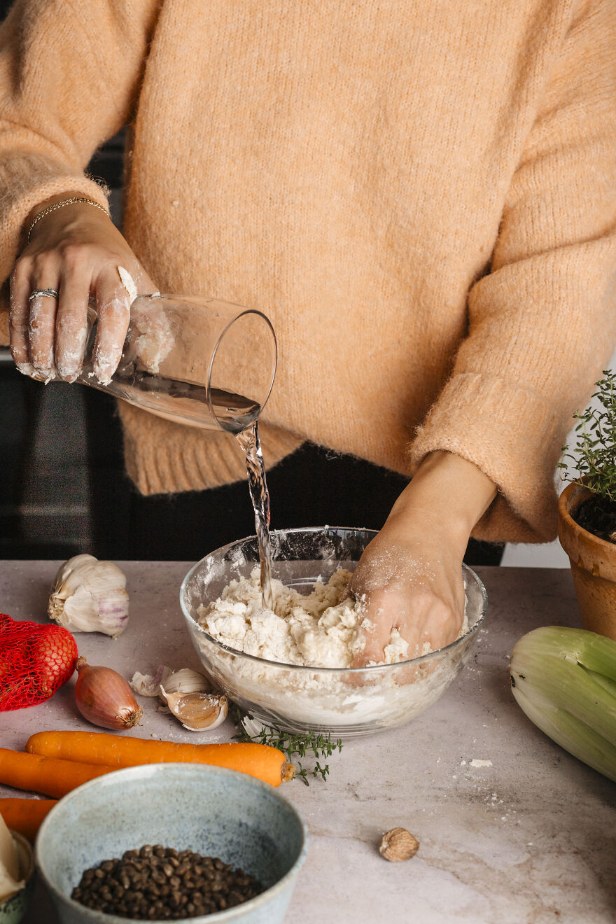 Person in cozy sweater pouring water into flour bowl preparing autumn pie recipe with fresh vegetables on table