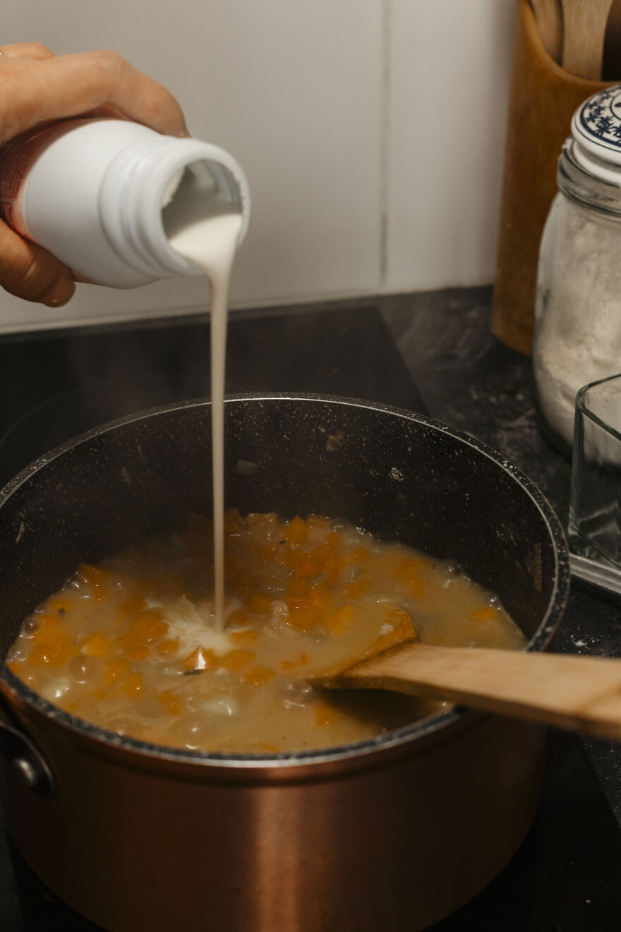 Pouring cream into a simmering pot of autumn pie filling with diced vegetables and a wooden spoon stirring.