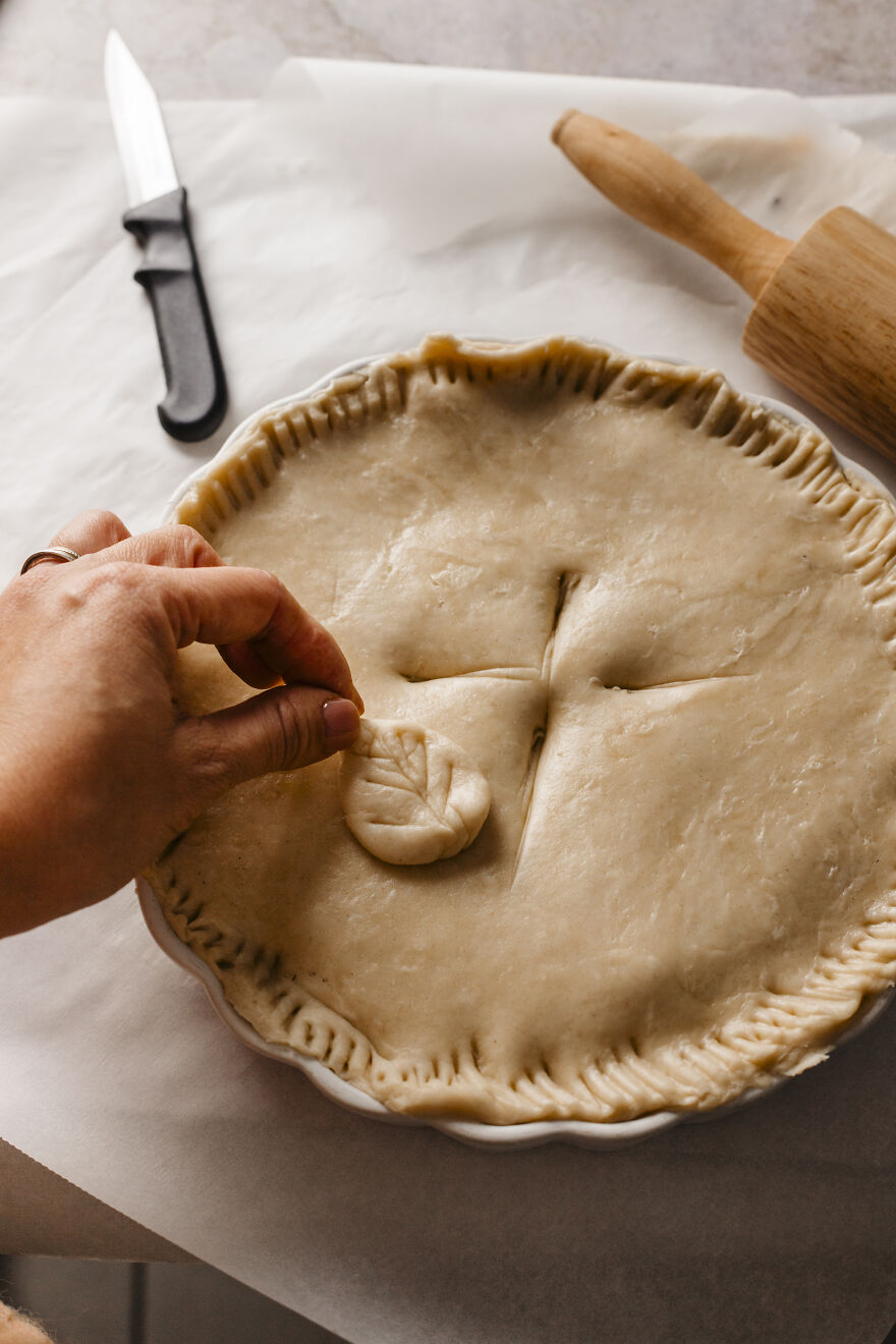 Hand placing a decorative leaf on an autumn pie crust, with rolling pin and knife nearby in preparation.