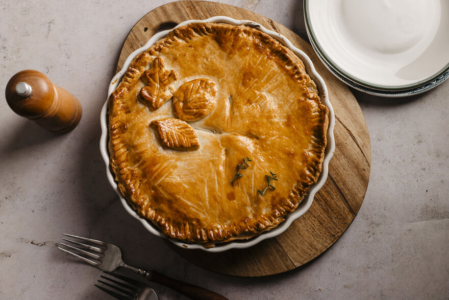 Golden autumn pie with decorative leaf crust, served on a wooden board beside plates and cutlery for a cozy meal.