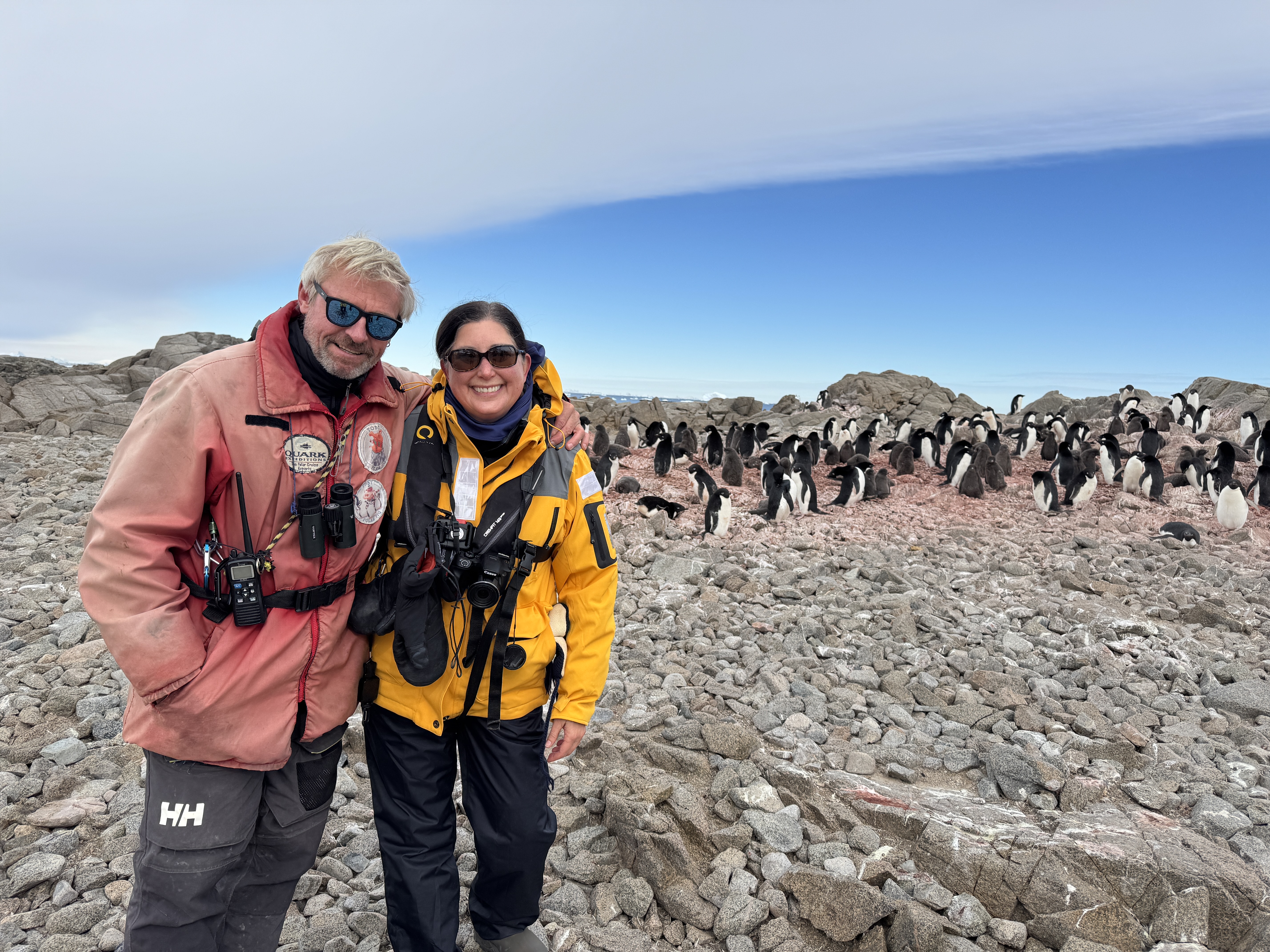 Two explorers in cold weather gear smiling on rocky land with a large group of penguins in Antarctica’s stunning landscape. Two explorers in cold weather gear smiling on rocky land with a large group of penguins in Antarctica’s stunning landscape.