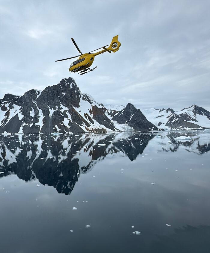 Yellow helicopter flying over reflective icy waters with snow-covered mountains in Antarctica adventure landscape. Yellow helicopter flying over reflective icy waters with snow-covered mountains in Antarctica adventure landscape.
