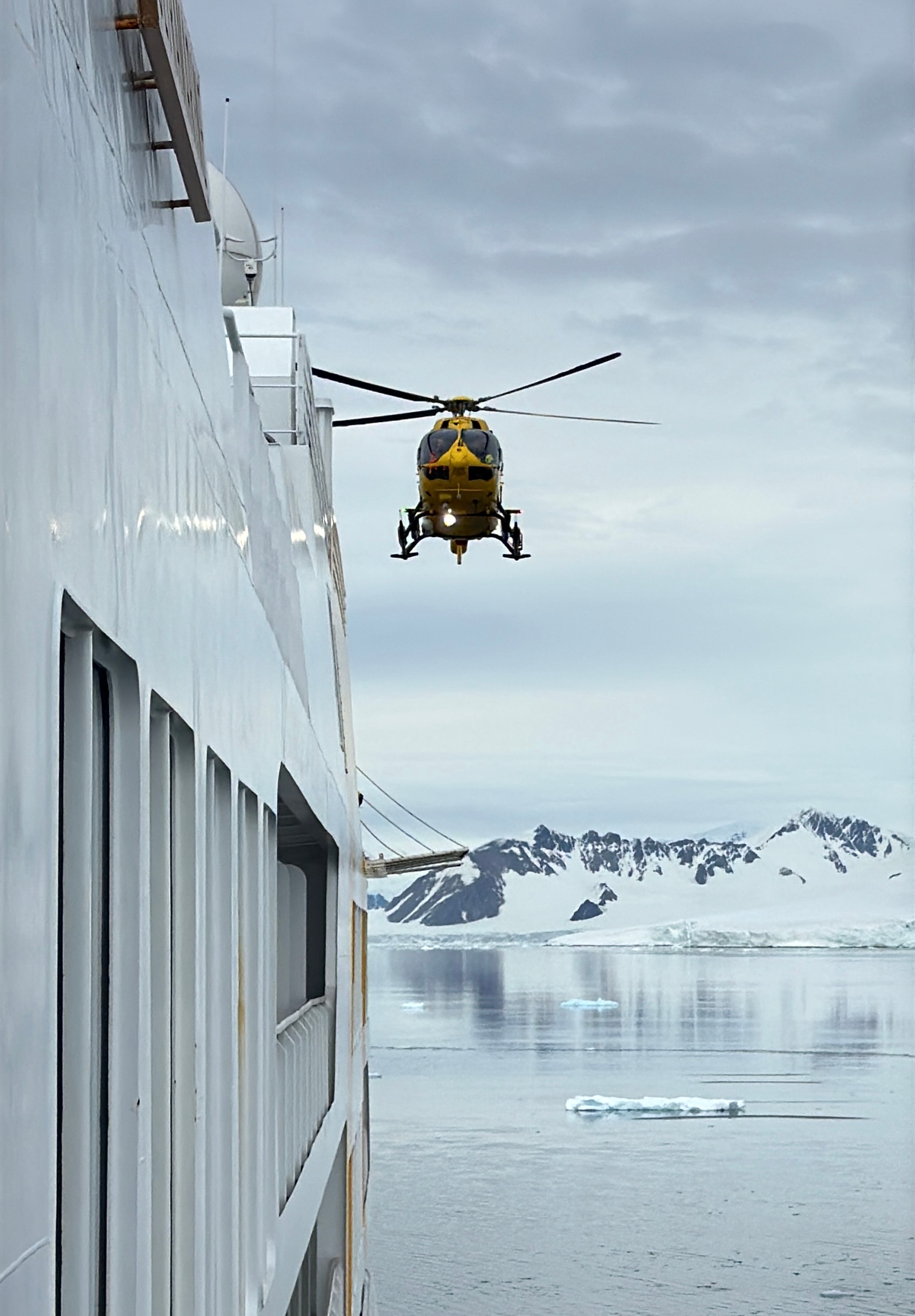 Yellow helicopter flying near a white ship with Antarctic icy mountains and calm water in the background. Yellow helicopter flying near a white ship with Antarctic icy mountains and calm water in the background.