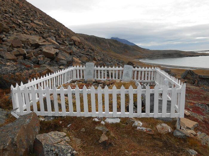 Remote location featuring a white picket fence grave site surrounded by rocky terrain and a distant coastline.