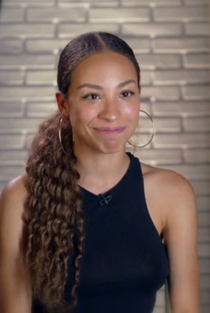 Young female singer with long curly ponytail and hoop earrings wearing a black sleeveless top during an interview session.